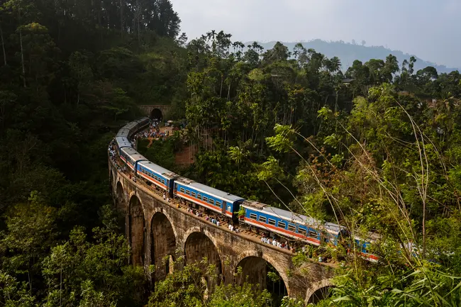 A train passes over a massive stone bridge that has nine arches and crosses a green jungle